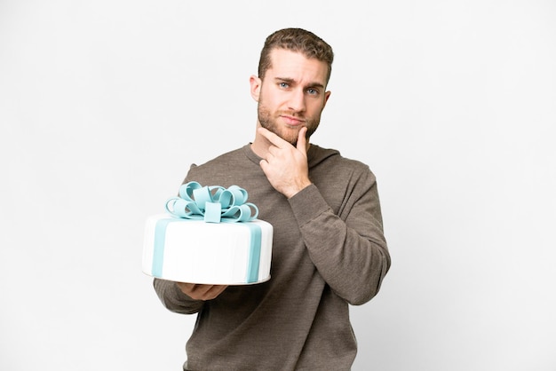 Young handsome blonde man with a big cake over isolated white background thinking