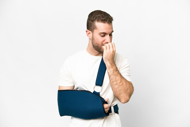 Young handsome blonde man with broken arm and wearing a sling over isolated white background having doubts