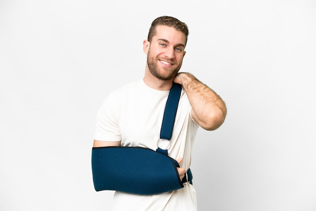 Young handsome blonde man with broken arm and wearing a sling over isolated white background laughing