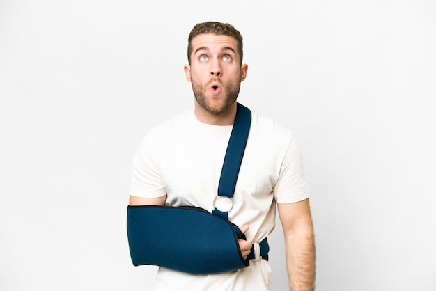 Young handsome blonde man with broken arm and wearing a sling over isolated white background looking up and with surprised expression