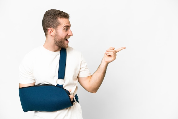 Young handsome blonde man with broken arm and wearing a sling over isolated white background pointing finger to the side and presenting a product