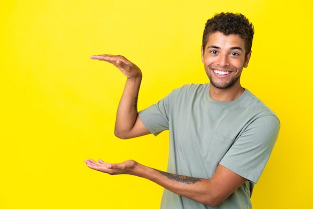 Young handsome brazilian man isolated on yellow background holding copyspace to insert an ad