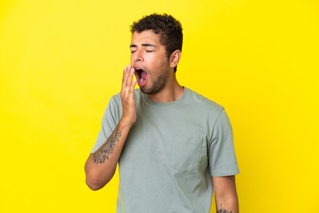 Young handsome Brazilian man isolated on yellow background yawning and covering wide open mouth with hand