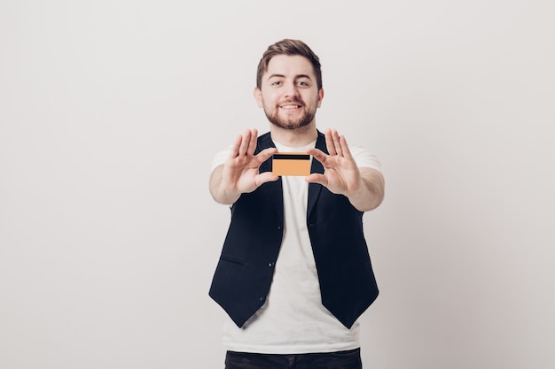 Giovane bell'uomo bruna con la barba in una camicia bianca e un gilet nero in possesso di una carta di credito in plastica e sorridente. concentrarsi sulla carta. luce soffusa