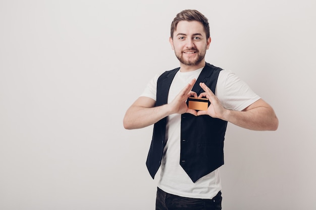 Young handsome brunette man with a beard in a white shirt and a black waistcoat holding a plastic credit card and smiling. soft light