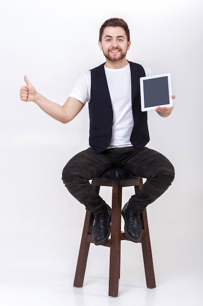 Young handsome brunette man with  beard in white shirt and  black waistcoat holding tablet and showing thumb up sign on gray background