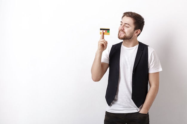 Young handsome brunette man with a beard in a white shirt and a black waistcoat showing a plastic credit card and smiling