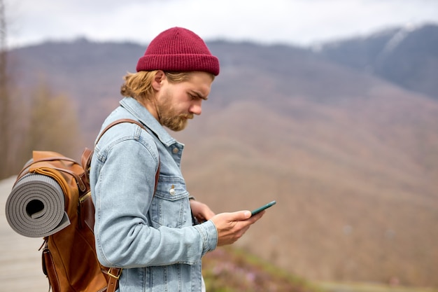 革の茶色のバックパック、秋のスタイル、旅行者、冒険、自由な精神で、スマートフォンを使用して野生の自然を旅する若いハンサムな残忍なひげを生やした男。コピースペース。側面図