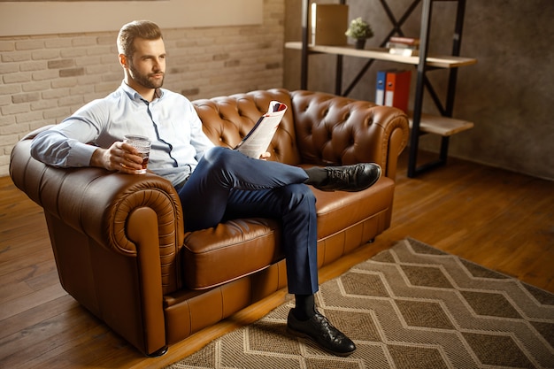 Young handsome buisnessman sitting on sofa and drink whiskey in his own office. He look straight with confidence. Guy hold journal. Attractive small smile.