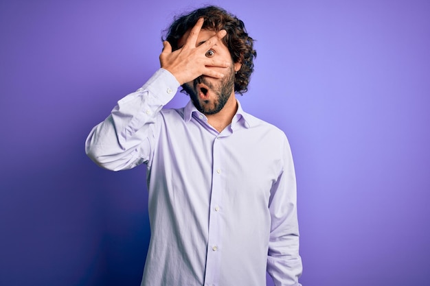 Young handsome business man with beard wearing shirt standing over purple background peeking in shock covering face and eyes with hand looking through fingers with embarrassed expression