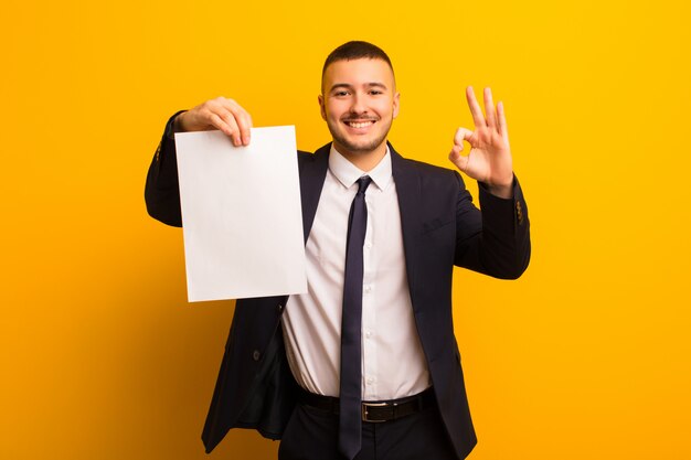 Young handsome businessman  against flat wall holding an empty piece of paper