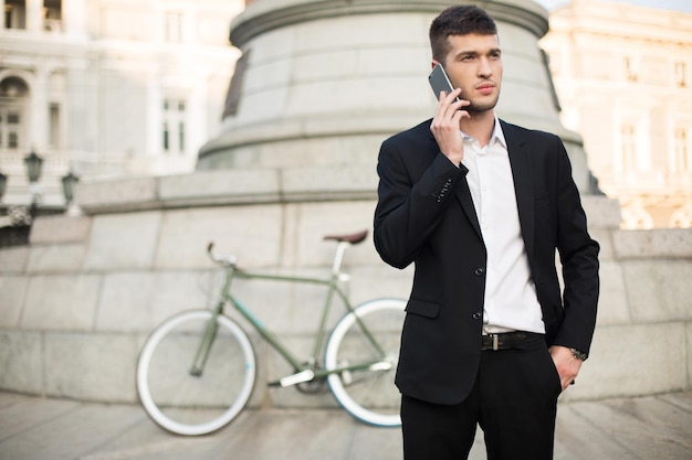 Young handsome businessman in classic black suit and white shirt talking on cellphone while thoughtfully looking aside with retro bicycle on background outdoor