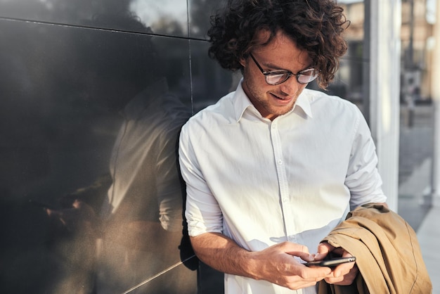 Young handsome businessman leaning on a wall while standing outdoors and using smartphone and free wireless Man with curly hair wearing white shirt and beige coat looking at his mobile phone