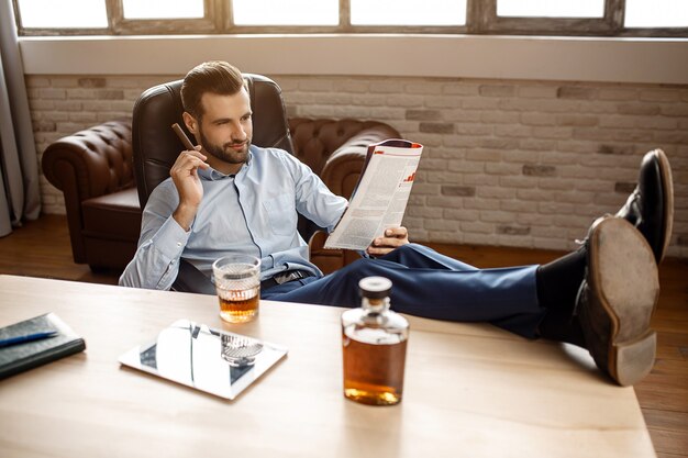 Young handsome businessman sit on chair and read journal in his own office. He hold cigar in hand and legs on table. Tablet with glass of whiskey and graphene stand together.