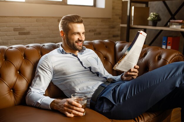 Young handsome businessman sit on sofa and read in his own office. He hold glass of whiskey in hand and smile. Positive and happy. Good-looking.