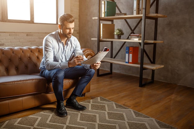 Young handsome businessman sit on sofa and read journal in his own office. He hold glass of whiskey in hand. Confident and sexy young man posing.