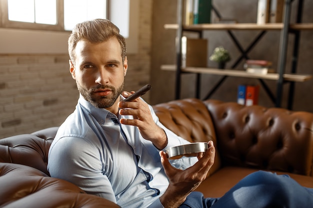 Young handsome businessman smoke cigar in his own office. He hold it and ash-pot. Guy look to side. Confidence. Daylight.