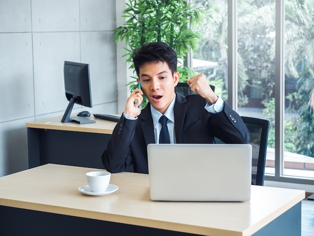 Young handsome businessman in suit raising fist with exciting and shocking while calling with his mobile phone and looking laptop computer on desk in office with winner feeling and celebrating.