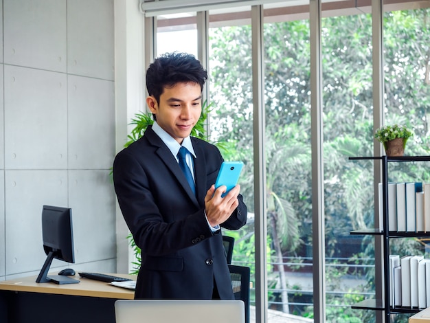 Giovane uomo d'affari bello in giacca e cravatta in piedi mentre si utilizza il telefono cellulare blu vicino alla scrivania con computer portatile, scaffale e finestra di vetro in ufficio con albero verde naturale.