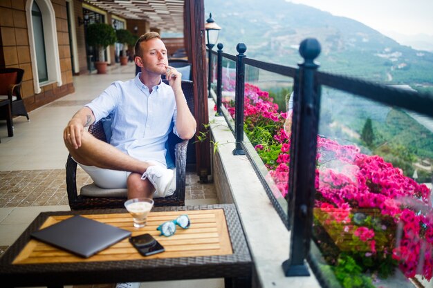 Young handsome businessman in a white shirt and shorts sits with a laptop in a cafe at the table. Work while resting