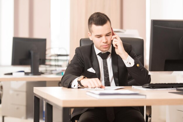 電話で話している現代のオフィスのデスクでコンピューターを使用して作業している若いハンサムなビジネスマン