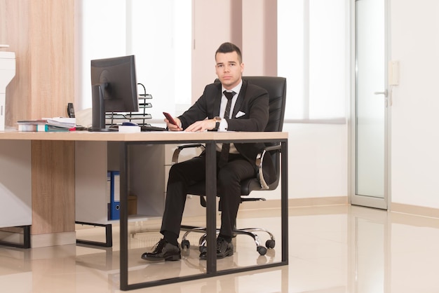Молодой красивый бизнесмен, работающий с компьютером за столом в современном офисе, разговаривает по телефону