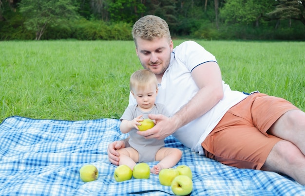 Young handsome caucasian blond father giving green apple to baby sitting on plaid in park on the picnic in summer. Healthy food, fruit for kids concept