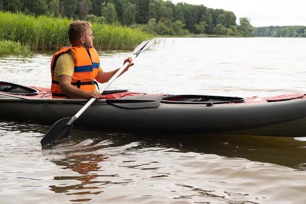 Foto giovane uomo biondo caucasico bello in canoa-kayak, barca a remi, indossando giubbotto di salvataggio arancione, giubbotto. idea di trascorrere del tempo all'aperto in natura, foresta verde, estate, sicurezza sul lago o sull'acqua, intrattenimento.