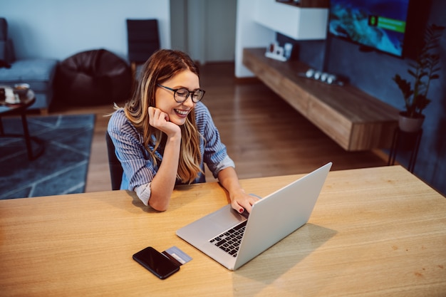 Young handsome caucasian brunette with eyeglasses sitting at dining table and using laptop.