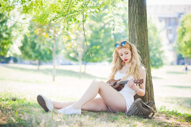 young handsome caucasian long blonde straight hair woman sitting on a city park playing with a ukulele