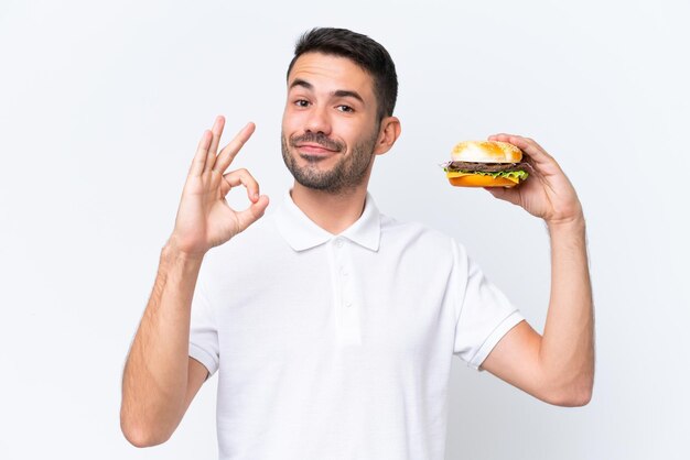 손가락으로 확인 표시를 보여주는 고립 된 배경 위에 햄버거를 들고 젊은 잘 생긴 백인 남자