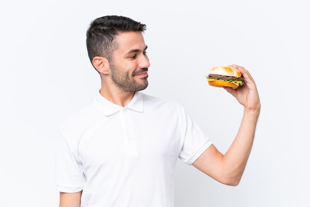 Young handsome caucasian man holding a burger over isolated background with happy expression