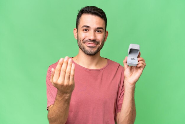 Young handsome caucasian man holding a engagement ring over isolated background inviting to come with hand Happy that you came