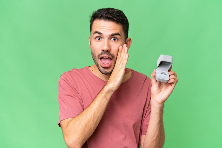 Photo young handsome caucasian man holding a engagement ring over isolated background whispering something