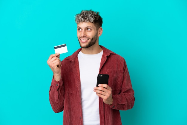 Young handsome caucasian man isolated on blue background buying with the mobile with a credit card while thinking