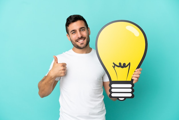 Young handsome caucasian man isolated on blue background holding a bulb icon with thumb up