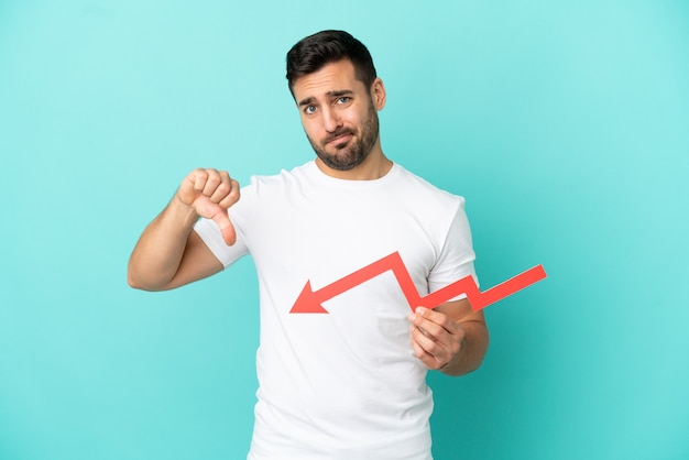 Young handsome caucasian man isolated on blue background holding a downward arrow and doing bad signal