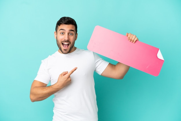 Young handsome caucasian man isolated on blue background holding an empty placard and pointing it