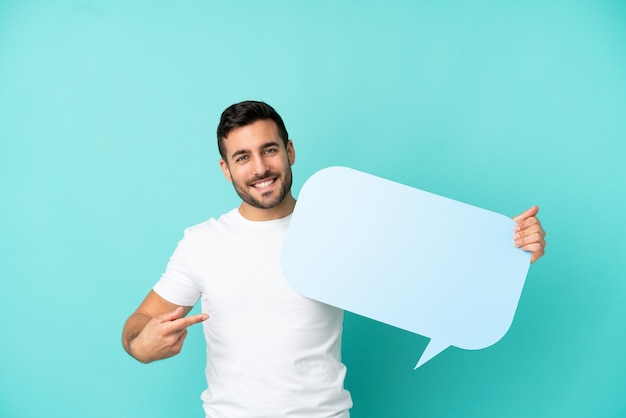 Young handsome caucasian man isolated on blue background holding an empty speech bubble and pointing it