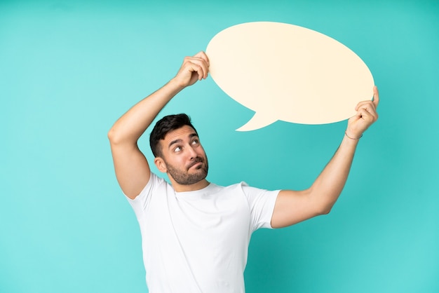 Young handsome caucasian man isolated on blue background holding an empty speech bubble and with sad expression