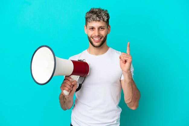 Young handsome caucasian man isolated on blue background holding a megaphone and pointing up a great idea