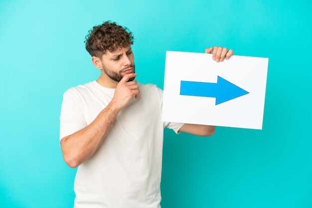 Young handsome caucasian man isolated on blue background holding a placard with arrow symbol and thinking