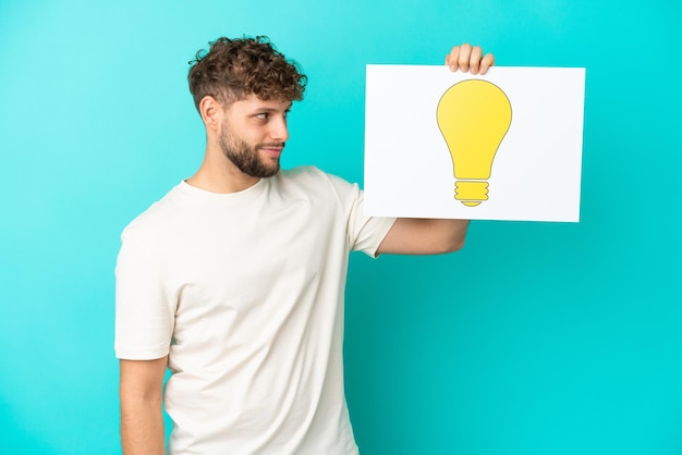 Young handsome caucasian man isolated on blue background holding a placard with bulb icon