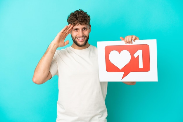 Young handsome caucasian man isolated on blue background holding a placard with Like icon with surprised expression