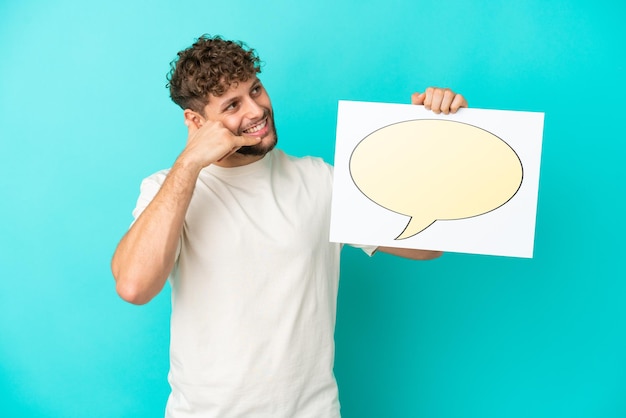 Young handsome caucasian man isolated on blue background holding a placard with speech bubble icon and doing phone gesture