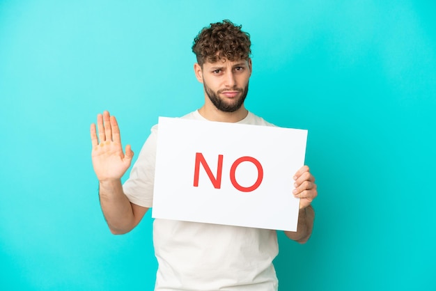 Young handsome caucasian man isolated on blue background holding a placard with text NO and doing stop sign