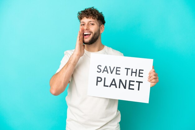 Молодой красивый кавказский мужчина, изолированные на синем фоне, держит плакат с текстом «Спасите планету» и кричит