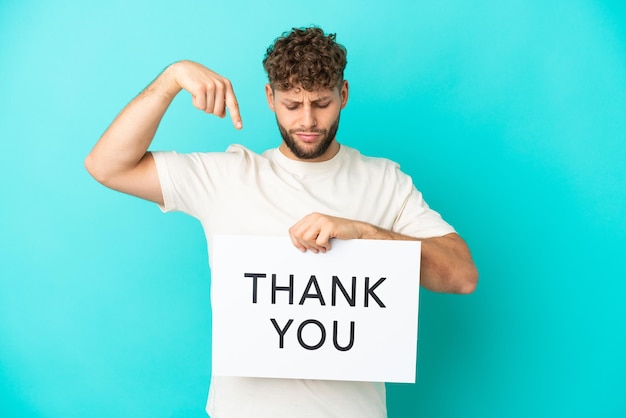 Young handsome caucasian man isolated on blue background holding a placard with text THANK YOU and  pointing it