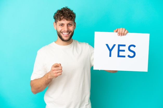 Young handsome caucasian man isolated on blue background holding a placard with text YES and pointing it