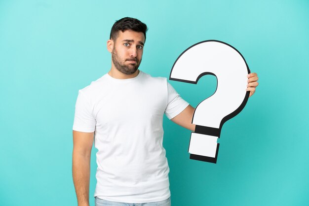 Young handsome caucasian man isolated on blue background holding a question mark icon and with sad expression